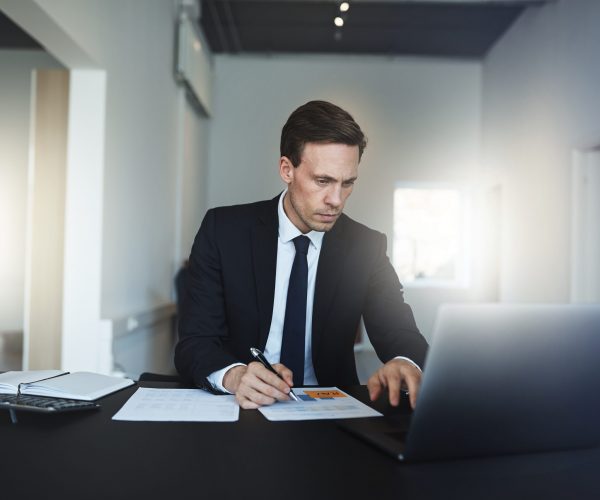 Businessman reading paperwork and using a laptop in an office Focused businessman going over paperwork and working on a laptop while sitting at his desk in a bright modern office